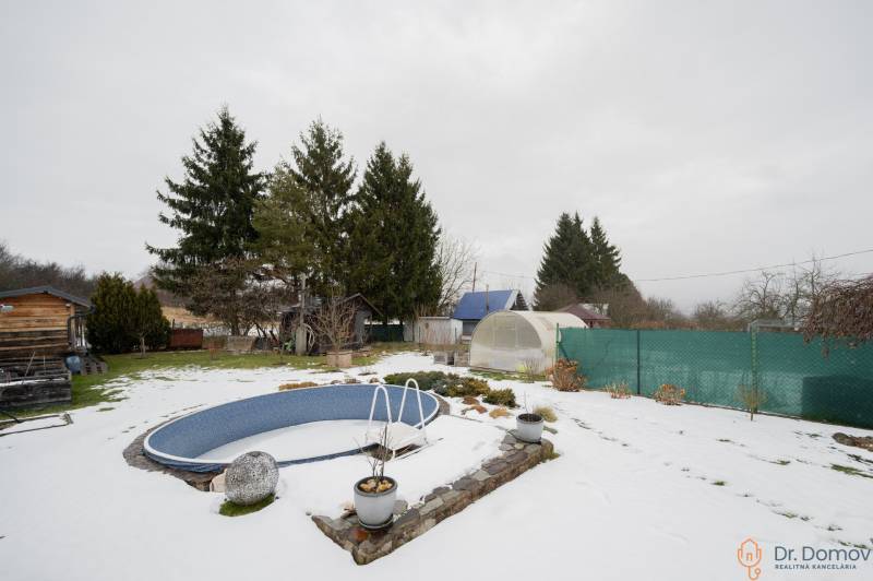 Ein verschneiter Garten bei einer Hütte in Handlová mit einem Pool, Gewächshäusern und Nadelbäumen.