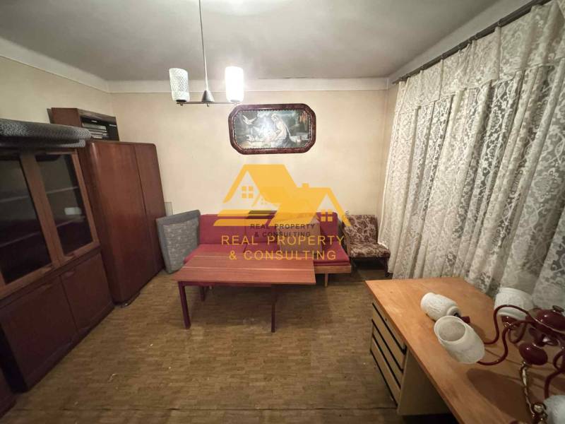 Ein Zimmer in einem Einfamilienhaus mit Möbeln, Vorhängen und einem Boden in Holzoptik.