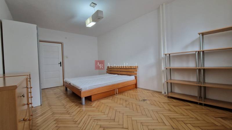 Schlafzimmer mit einem einfachen Bett, Regalen und einem Boden mit Holzdekor in einer 2-Zimmer-Wohnung.