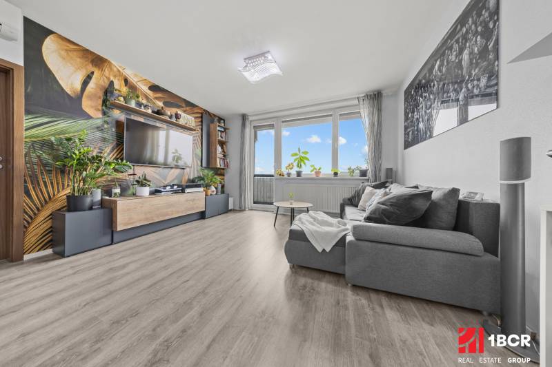 Wohnzimmer mit einem bequemen Sofa, einer Tapete mit Pflanzenmotiv und einem Boden in Holzoptik.