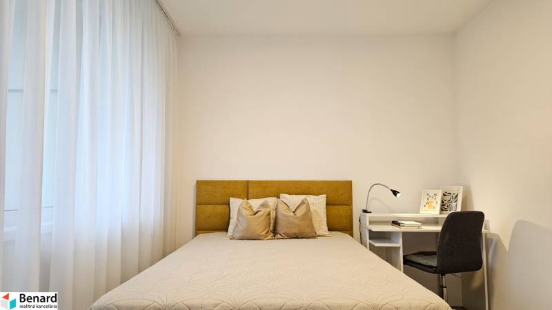 Schlafzimmer in einer Einzimmerwohnung mit hellem Bett, Vorhängen und einem weißen Tisch mit Lampe.