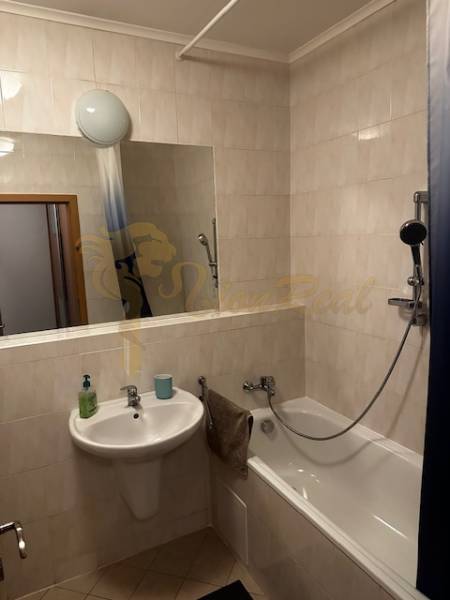 Badezimmer in einer Einzimmerwohnung mit Badewanne, Waschbecken und Spiegel, helle Fliesen.