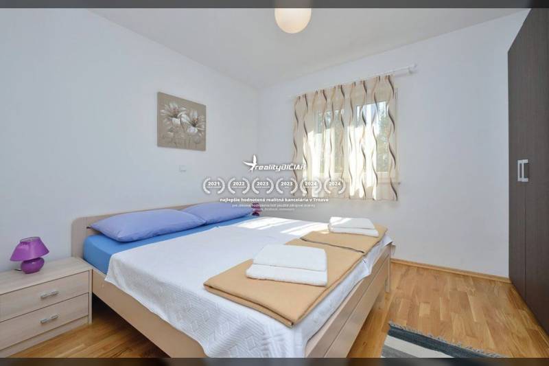 Schlafzimmer in einer 2-Zimmer-Wohnung mit Bett, Holzboden und Fenster mit zarten Vorhängen.