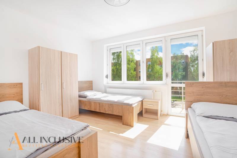 Ein heller Raum mit drei Betten, weißen Wänden, Schränken und großen Fenstern.