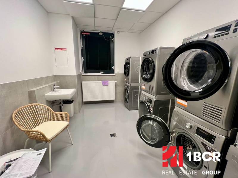 Waschküche mit Waschmaschinen, Trocknern, Waschbecken und Stuhl in einer Zweizimmerwohnung.