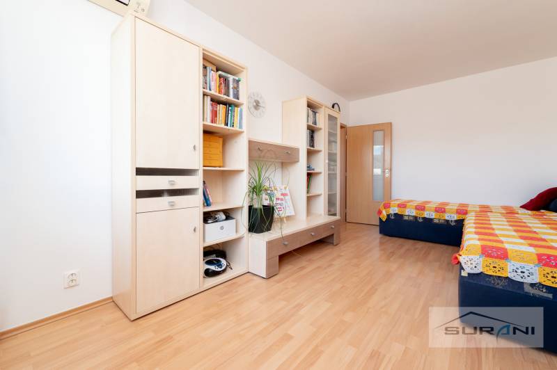 Ein Zimmer mit einem Holzboden enthält Möbel, Regale, ein Doppelbett und eine bunte Decke.