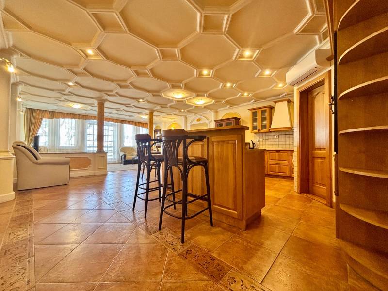 Innenraum einer 4-Zimmer-Wohnung mit dekorativer Decke, Holzboden, Kücheninsel und Stühlen.