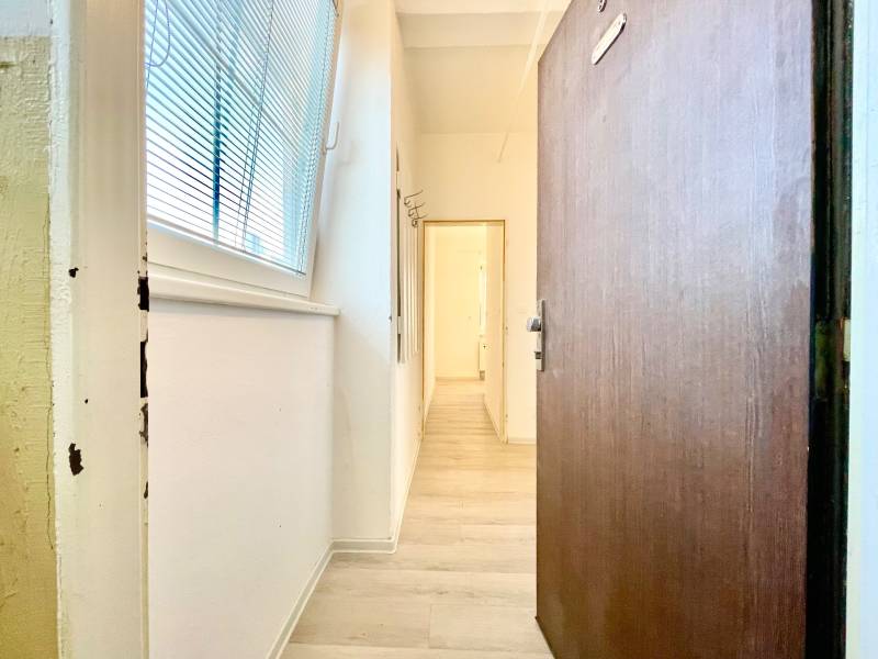 Ein Flur mit einem Boden in Holzoptik, weißen Wänden, einem großen Fenster mit Jalousien.