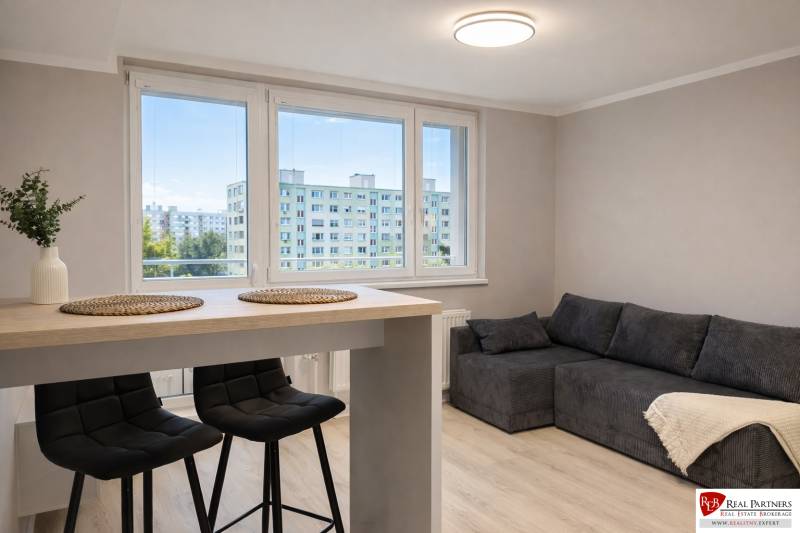 Obývací Zimmer in einer Zweizimmerwohnung mit Sofa, Bartisch und Stühlen.