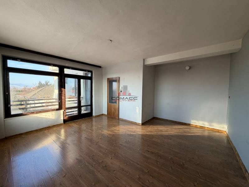 Ein Zimmer in einem Einfamilienhaus mit einem Boden in Holzoptik, einem großen Fenster und einer Tür.