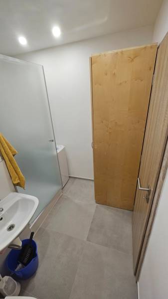 Badezimmer in einem Studio-Apartment mit Waschbecken, Duschkabine und Schrank auf hellem Fliesenboden.