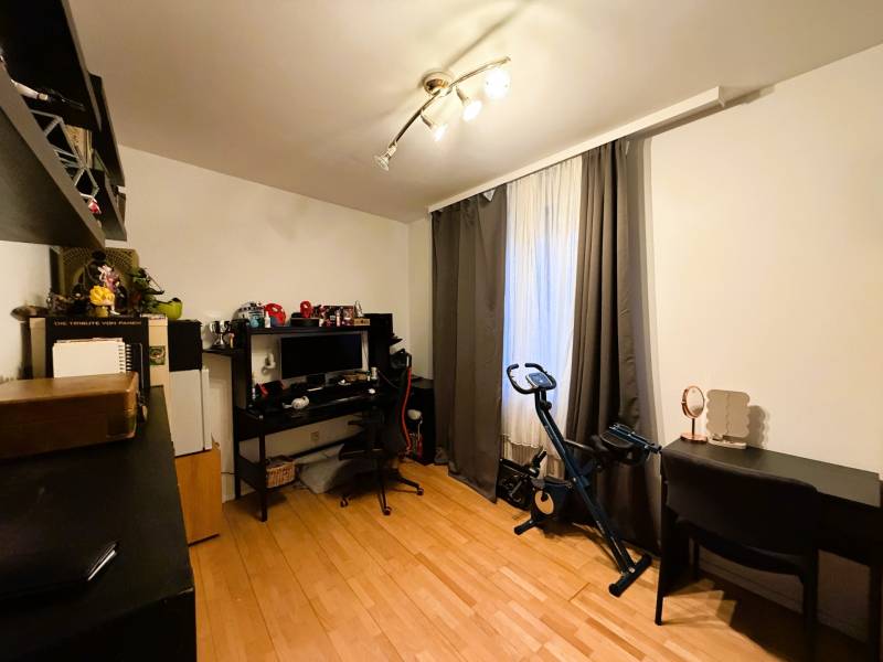 Ein Zimmer mit einem Holzboden, einem Schreibtisch, einem Computer, einem Heimtrainer, Regalen.