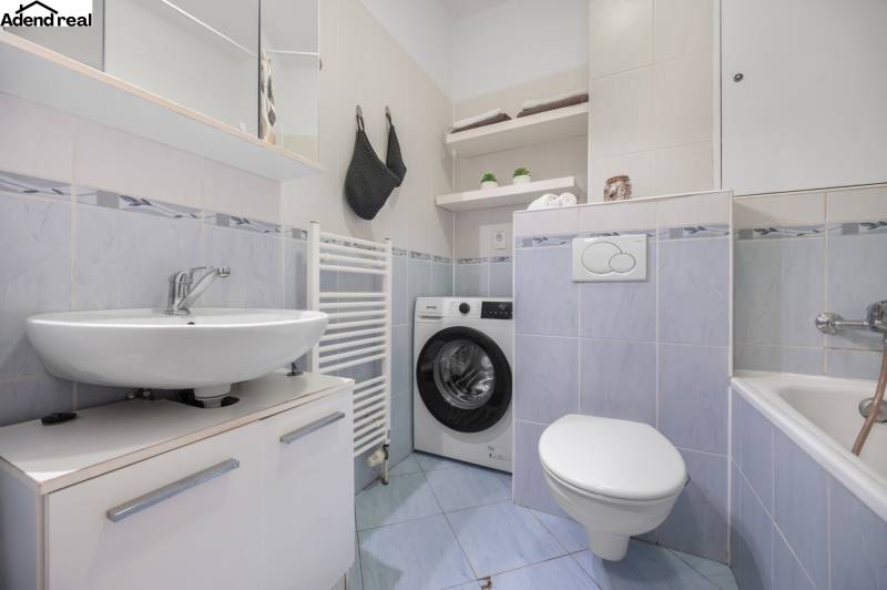 Badezimmer in einer Einzimmerwohnung mit Waschmaschine, Waschbecken und Badewanne, hellblaue Fliesen.