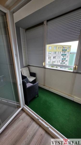 Balkon in einer 3-Zimmer-Wohnung mit einem Rattansessel und einem grünen Teppich.