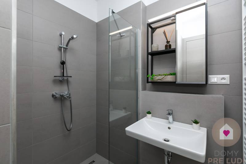 Graues Badezimmer mit Dusche, Waschbecken und großem Spiegel in einer Einzimmerwohnung.