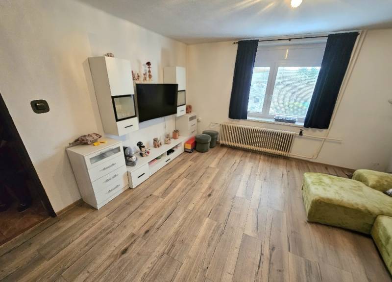 Wohnzimmer in einem Einfamilienhaus mit Fernseher, weißen Möbeln und einem Boden in Holzoptik.