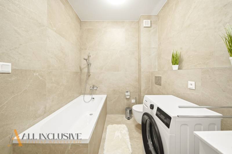 Ein Badezimmer mit hellen Fliesen, einer Badewanne, einer Waschmaschine und dekorativen Pflanzen in einer 4-Zimmer-Wohnung.