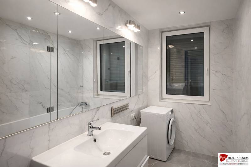 Badezimmer in einer 2-Zimmer-Wohnung mit Badewanne, Waschbecken und Waschmaschine, Fliesen in Marmoroptik.