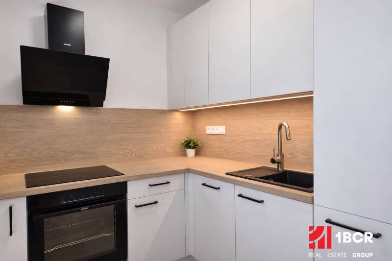 Küche in einer 2-Zimmer-Wohnung mit Holzdekor, weißen Schränken und einem schwarzen Spülbecken.