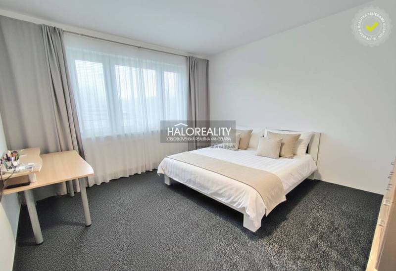 Schlafzimmer mit hellem Bett und Tisch in einer 4-Zimmer-Wohnung, weiße Wände und grauer Teppich.