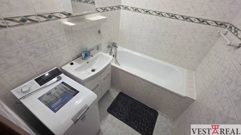 Badezimmer mit Badewanne und Waschmaschine in einer Einzimmerwohnung, helle Fliesen und Stauraum.