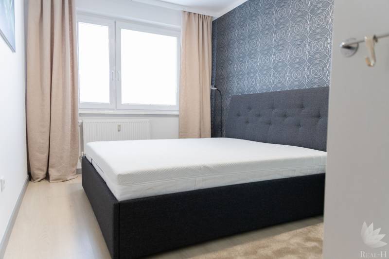 Schlafzimmer in einer 4-Zimmer-Wohnung mit Bett, Tapete, Fenstern und Vorhängen, Boden mit Holzdekor.