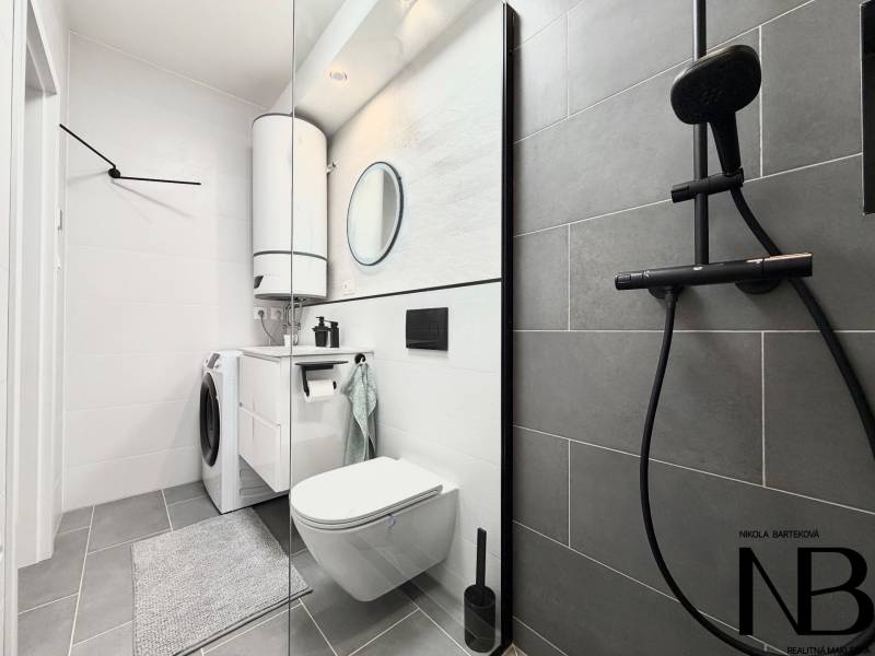 Badezimmer in einer Einzimmerwohnung mit Waschbecken, Dusche und Waschmaschine auf hellem Fliesenboden.