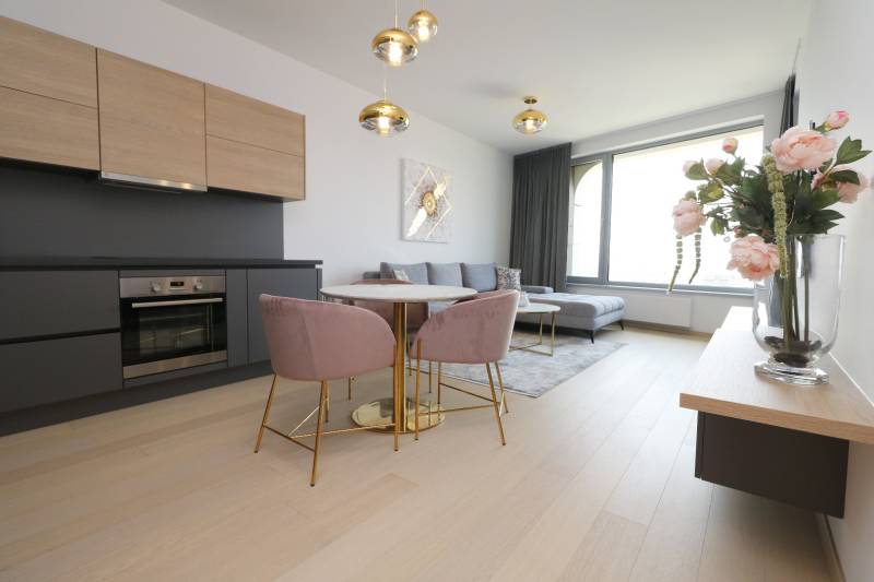 Wohnzimmer mit Küche, Boden mit Holzdekor, helle Couch, Esstisch, Blumen.