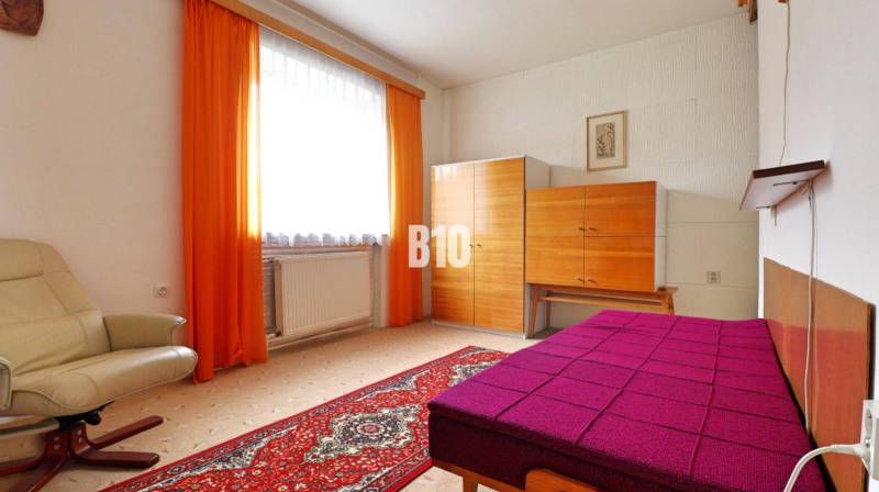 Ein Zimmer mit Teppichboden, einem cremefarbenen Sessel, einem bunten Bett und einem Schrank in einem Einfamilienhaus.