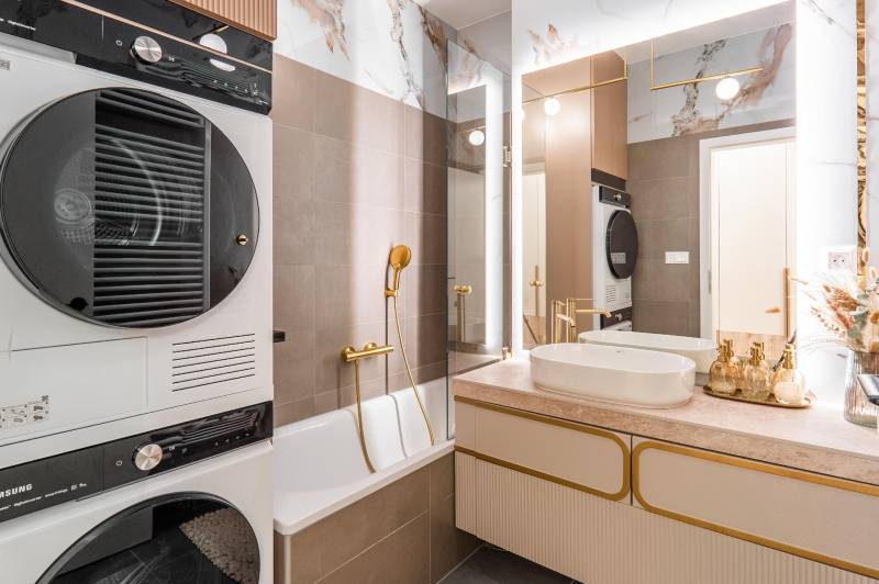 Badezimmer in einer 3-Zimmer-Wohnung mit Badewanne, Waschbecken, Trockner und eleganten goldenen Accessoires.