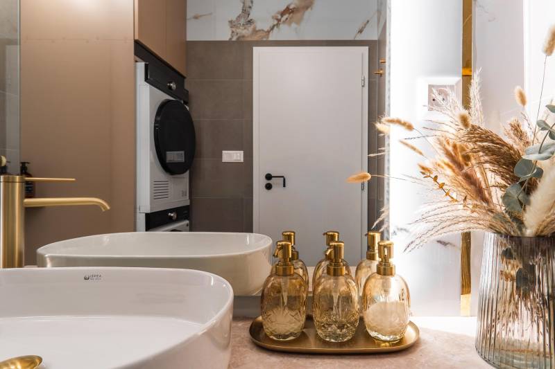 Badezimmer mit Waschbecken, goldenen Spendern und dekorativer natürlicher Dekoration in einer 3-Zimmer-Wohnung.