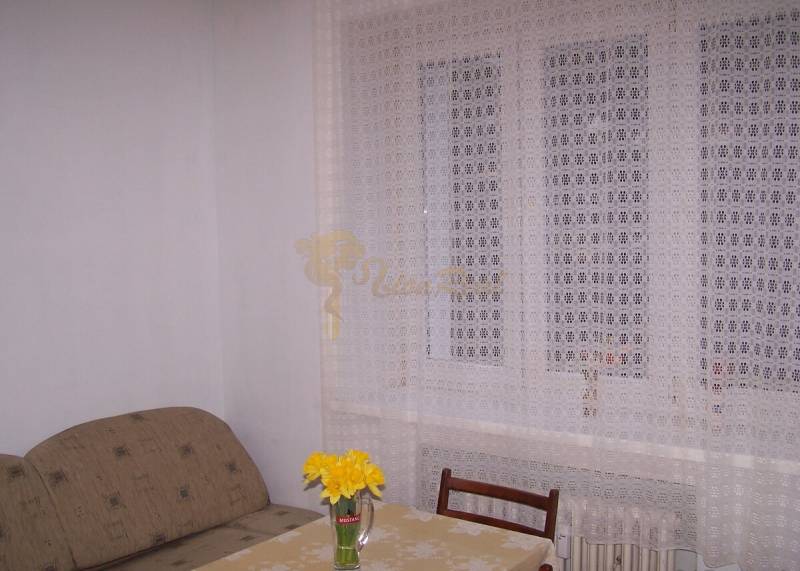 Ein Zimmer mit einem Sofa, einem Tisch, einem Stuhl und einem Vorhang, auf dem Tisch eine Vase mit Blumen.