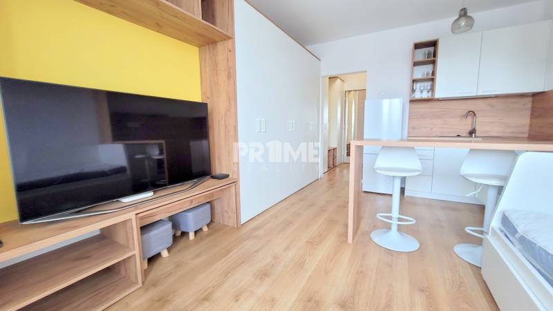 Einzimmerwohnung mit Holzboden, Fernseher, weißer Kücheneinrichtung und Barhockern.