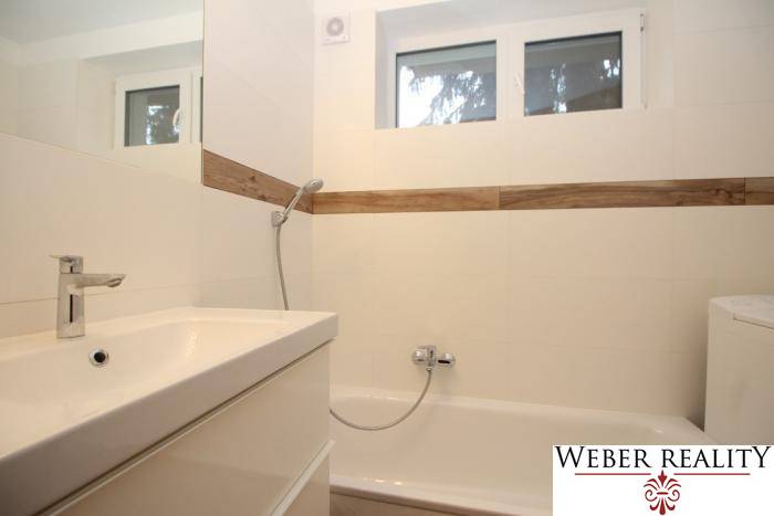 Badezimmer mit Badewanne und Waschbecken in einer Einzimmerwohnung, weiße Fliesen, Holzelemente.