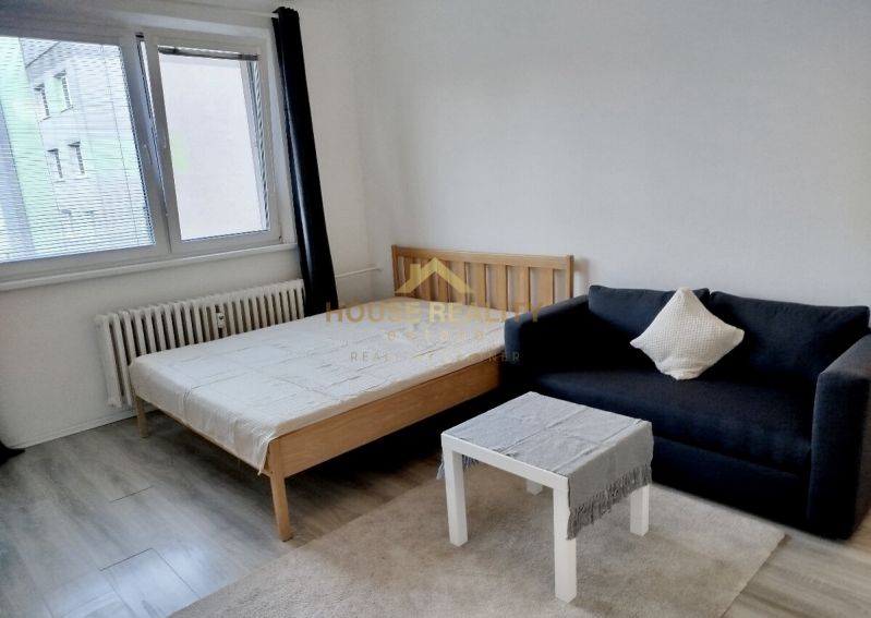 Izba s posteľou, čiernou pohovkou a stolíkom, podlaha s dreveným dekorom.