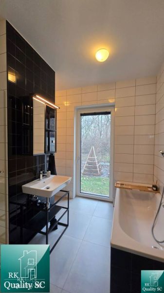 Badezimmer mit Badewanne und Waschbecken in einem Einfamilienhaus, weiße und schwarze Fliesen, Fenster mit Aussicht.