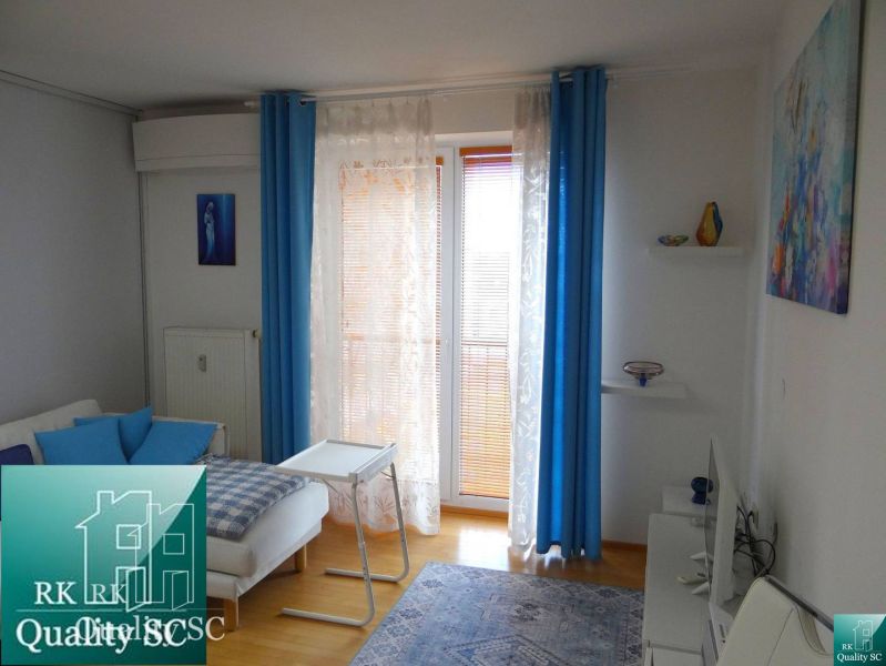 Ein Zimmer in einer Einzimmerwohnung mit weißen Wänden, einem Boden in Holzoptik und blauen Vorhängen.