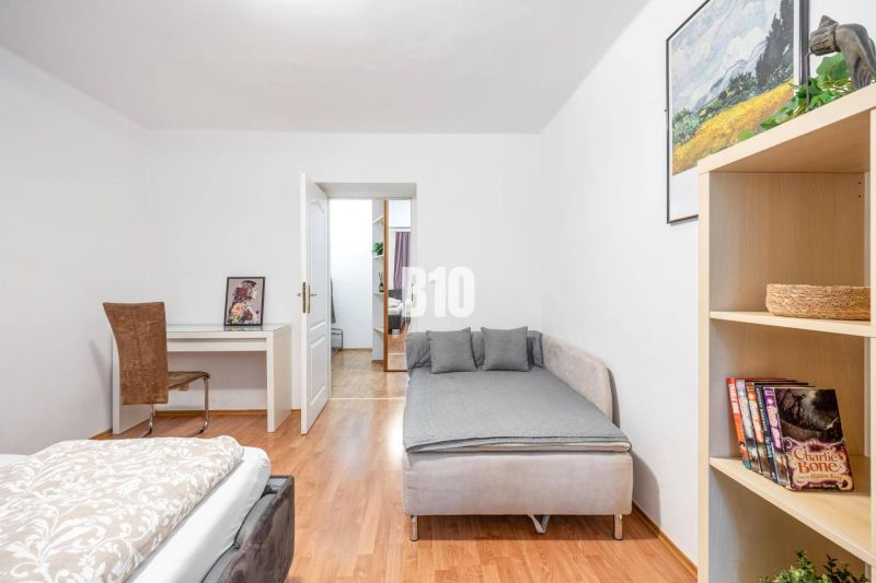 Helles Zimmer mit Bett, Tisch, Regal und Boden in Holzoptik.