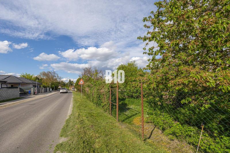 Straße in Dunajská Lužná mit Grundstücken für Wohnzwecke, umgeben von Grünflächen und einem Zaun.