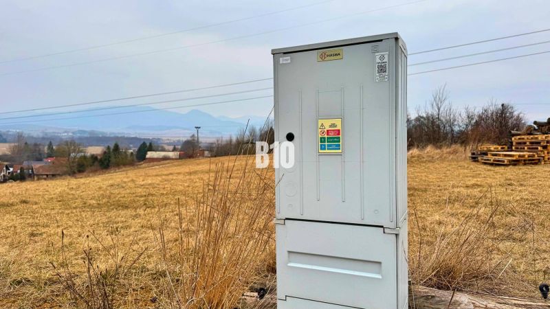 Ein elektrischer Schrank auf einer Wiese, im Hintergrund Hügel und ländliche Bebauung.