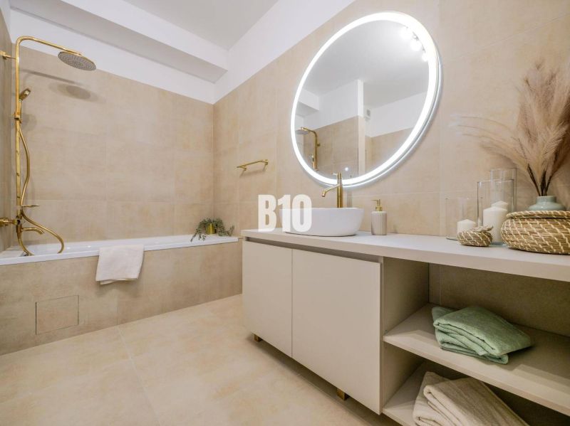 Badezimmer in einer 2-Zimmer-Wohnung mit Badewanne, rundem Spiegel und beigen Fliesen.