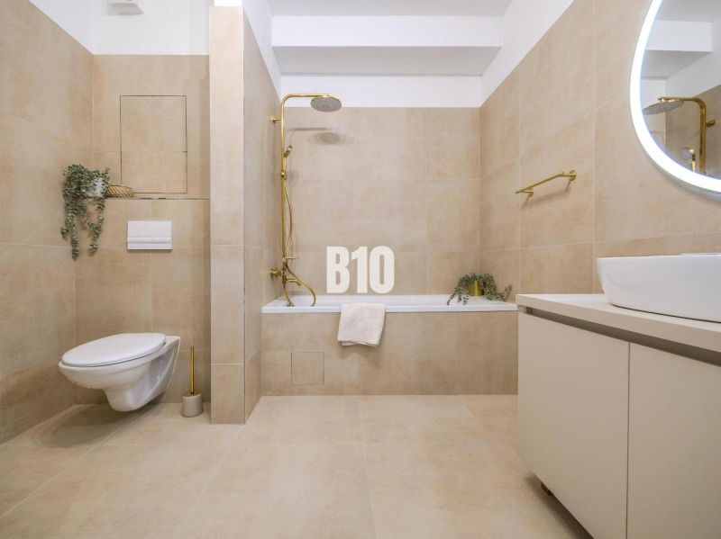 Badezimmer in einer 2-Zimmer-Wohnung mit beigen Fliesen, eingebauter Badewanne und hängender Toilette.