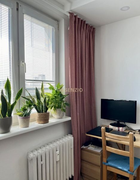 Ein Zimmer mit einem Tisch, einem Monitor, Pflanzen auf der Fensterbank und einem bordeauxroten Vorhang in einer Zweizimmerwohnung.