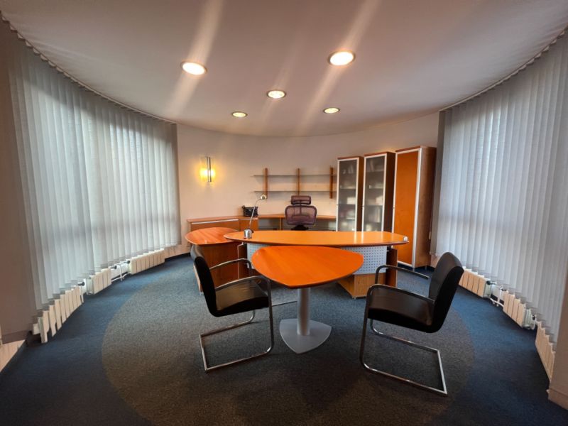 Ein Büro mit einem Tisch in Holzoptik, Regalen und beleuchteten vertikalen Jalousien.