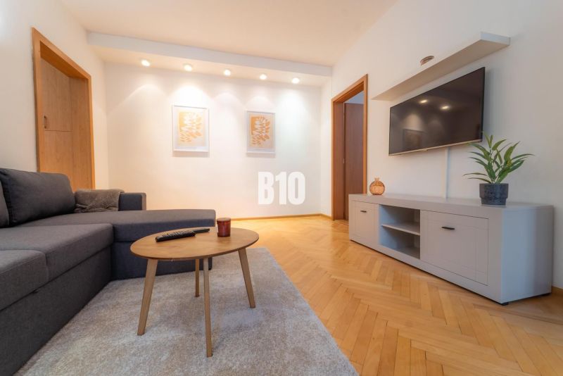 Wohnzimmer mit einem Fernseher an der Wand, einem grauen Sofa und einem Boden in Holzoptik.