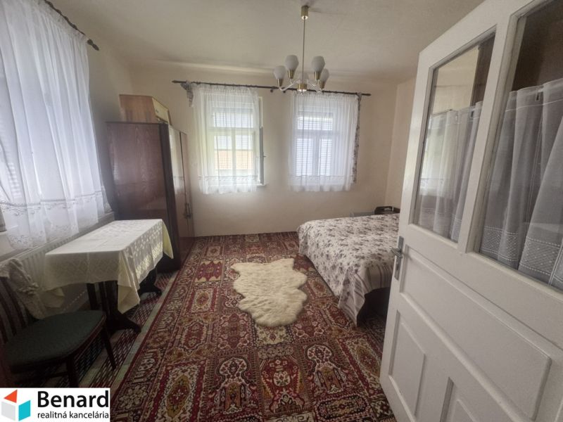 Ein Zimmer in einem Einfamilienhaus mit Teppich, Bett, Tisch und Vorhängen an den Fenstern.