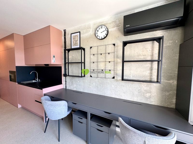 Ein Studio mit einem Boden in Holzoptik, einer rosa Küche und grauen Arbeitsstühlen.