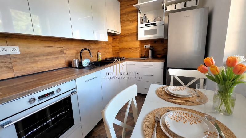 Küche in einer 2-Zimmer-Wohnung mit weißen Schränken, Holzboden und einem Tisch mit Blumen.