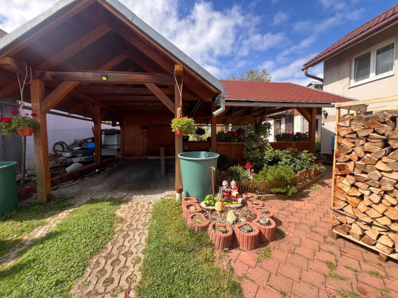 Ein Einfamilienhaus in Bacúch mit einem Unterstand, einem Garten und gelagertem Holz, an einem sonnigen Tag.