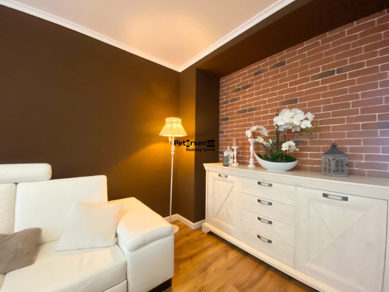 Wohnzimmer einer Zweizimmerwohnung mit bequemer Couch, Ziegelverkleidung und Boden in Holzoptik.